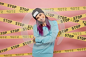 Smiling girl with purple hair tips dressed in blue sweatshirt and black hat stands on the background of pink wall with