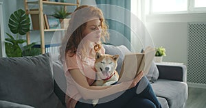 Smiling girl reading book at home sitting on sofa and petting cute puppy