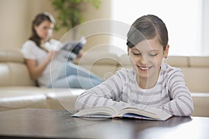 Smiling girl reading book with mother in background at home