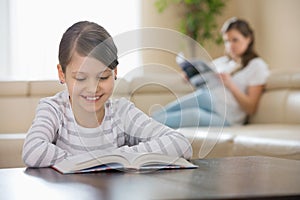 Smiling girl reading book with mother in background at home