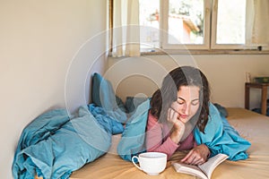 Smiling girl reads a book and drinks coffee while lying in bed