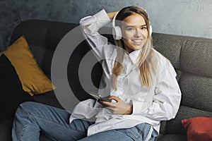 Smiling girl relaxing at home, listening to music in headphones. Female using smartphone with online services