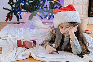 Smiling girl in Santa hat writing letter for gifts to Santa Claus. Christmas miracle wish list. Child lies at decorated