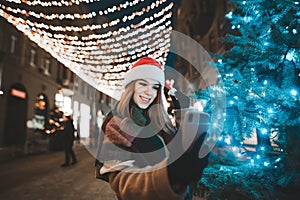 A smiling girl in Santa`s hat stands near a Christmas tree on a New Year`s Eve street decoration, makes a selfie on a smart phon