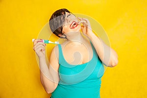 Smiling girl with short hair and a curvy figure stands with a toothbrush in her hands on yellow solid studio background
