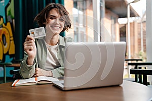 Smiling girl showing credit card while working with laptop in cafe