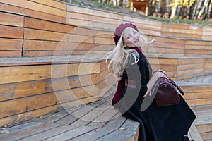 A smiling girl sits on a summer theater bench, made of wood in a burgundy coat and biret, an adult looks at the camera