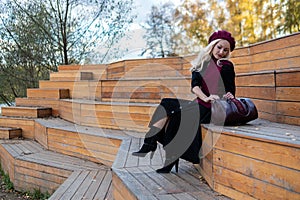 A smiling girl sits on a summer theater bench, made of wood in a burgundy coat and biret, smiles at the camera, in the