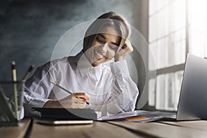 Smiling girl sitting at desk, making notes with pen in copybook. Female student studying in front of laptop