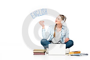 Smiling girl sitting on floor in headphones with speech bubble with english lettering near laptop, books and copybooks