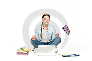 Smiling girl sitting on floor with laptop, books and copybooks and holding uk flag