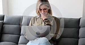 Smiling girl is sitting, relaxing on sofa and using modern laptop