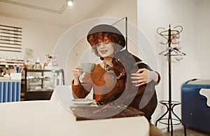 Smiling girl in stylish clothes and hat spends time in cafe with dog, happy drinking coffee. Hipster girl sitting in a cramped