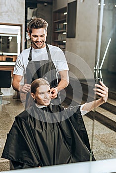 Smiling girl taking selfie with smartphone while hairstylist doing haircut