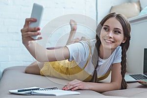 smiling girl taking selfie on smartphone while lying on sofa with copybook