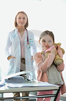 Smiling girl with teddybear standing against doctor at hospital