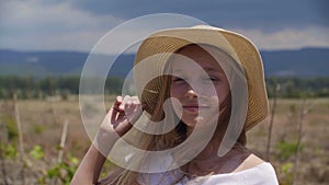 Smiling girl teenager in hat looking to camera on summer field and mountain landscape. Pretty teen girl posing front