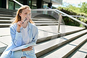 Smiling girl university student holding notebooks sitting on stairs outdoor.