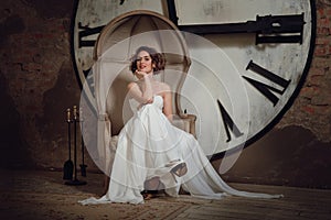 A smiling girl in a wedding dress in strange chair. The bride in a chair on the background of clocks and fireplace tool set.