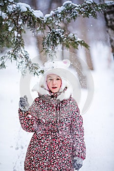 Smiling girl with white fur hat like a cat playing with snow. Winter snowy background and gteen trees