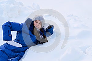 smiling girl in winter blue coat is lying on snow with her head propped on her hand. Girl against background of winter snowdrifts