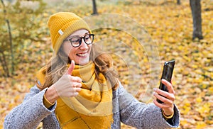 Smiling girl in a yellow hat and scarf showing thumb up takes selfie in the autumn park