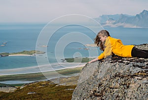 Smiling girl in yellow jacket and sunglasses lies on stone in on the edge of rocks in mountains and looking to fjord