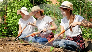 Smiling girls with mother cultivating soil and digging hole in garden bed for planting new vegetable seedlings and