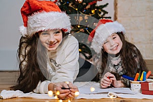 Smiling girls in Santa hat writing letter for gifts to Santa Claus. Christmas miracle wish list. Child lies at decorated