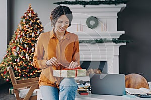 Smiling glad italian female tying red ribbon on Christmas present while sitting at home office
