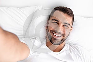 Smiling glad millennial muscular caucasian man waking up taking selfie on white bed on soft pillow in bedroom