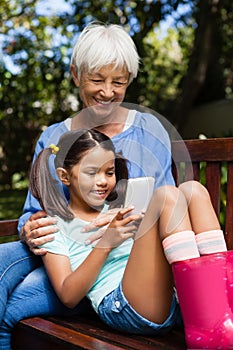 Smiling grandmother sitting with granddaughter using mobile phone on wooden bench