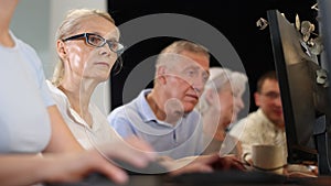 Smiling gray-haired elderly woman in glasses participating in group lesson on computer literacy basics, demonstrating