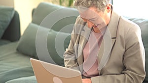 Smiling gray-haired lady in a business suit is working with a laptop at home alone while sitting on the couch. Remote