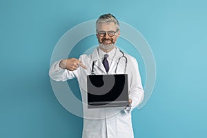 Smiling grey-haired caucasian doctor showing laptop with blank screen