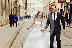 Smiling groom holds bride`s hand while they run along the tramwa