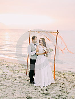 Smiling groom is hugging the bride back near the wedding arch placed on the beach during the sunset.