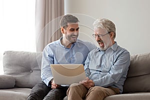 Smiling grown son and senior dad laugh using laptop together