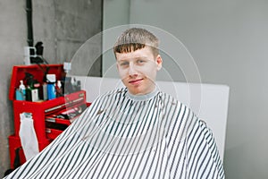 Smiling guy in a peignoir sits in a barber shop`s chair, looks into the camera and smiles against a light wall background. Guy