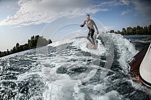 Smiling guy rides surfboard on the waves behind boat.