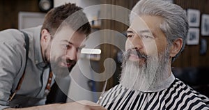Smiling hairdresser cutting beard and smiling looking at male client in barbershop