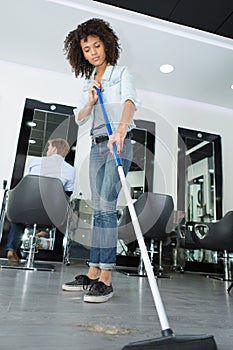 Smiling hairstylist sweeping hair clippings on floor in salon