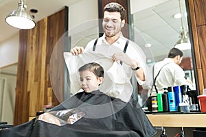 Smiling Hairstylist Wiping Head Of Little Client In Barber Shop