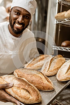 smiling handsome african american baker with tray of fresh loaves of bread