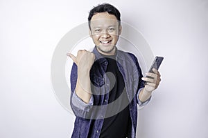 Smiling handsome Asian man pointing finger sideward to empty space while holding his phone  white background