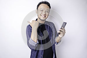 Smiling handsome Asian man pointing finger sideward to empty space while holding his phone  white background