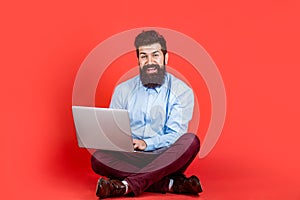 Smiling handsome bearded man worker laptop. Happy young man sitting on the floor with and using laptop computer on red