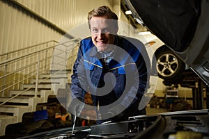 Smiling handsome Caucasian man, professional mechanic, auto technician repairing car engine at car service workshop