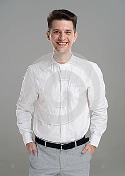 Smiling handsome confident young man standing in a white shirt on grey background with hands on pockets