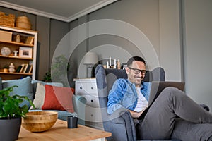 Smiling handsome freelancer checking e-mails over wireless computer while relaxing on armchair at home. Young male entrepreneur